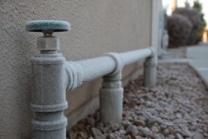 A frost-covered outdoor water pipe runs along a building wall above rocky ground, indicating cold weather conditions.