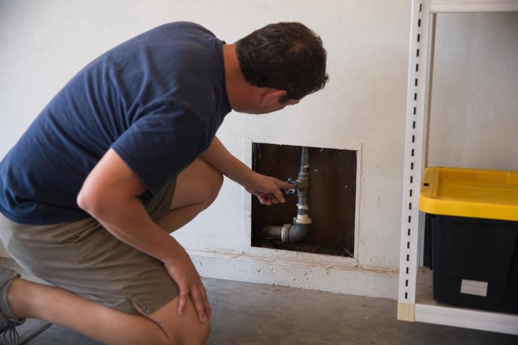 A person kneels by an open wall access panel, inspecting or adjusting pipes inside. Nearby, there is a white shelving unit with a black and yellow storage bin.