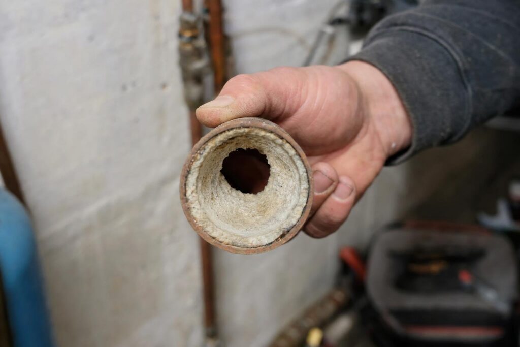A person holds a pipe with a thick layer of white mineral buildup inside, showing severe clogging. The background is a workshop or basement setting.