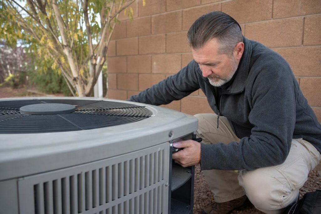 A man kneels outdoors next to a large air conditioning unit, inspecting or repairing it with a tool. He wears a dark jacket and light pants, and the scene is set next to a brick wall and some trees.